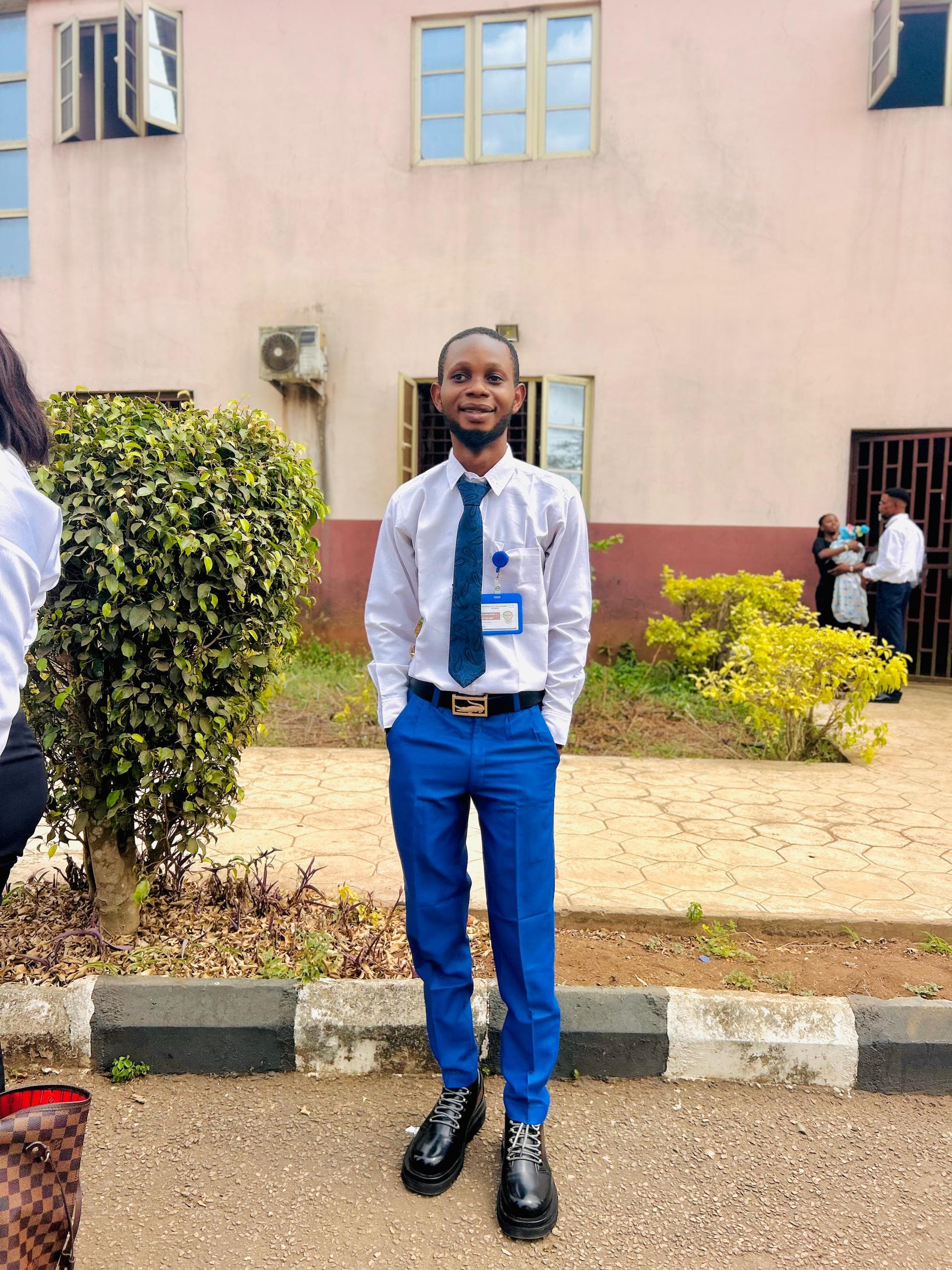 Smiling man in a white shirt, blue tie, and blue trousers posing outside a building.